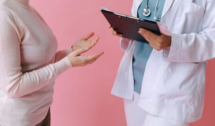 Kobieta w jasnoróżowym golfie rozmawia z lekarką w białym fartuchu i ze stetoskopem, która trzyma tablet. Różowe tło. 